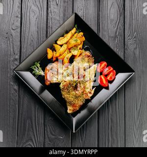 Gebratene Hähnchenschenkel mit gebackenen Kartoffeln und Kirschtomaten auf schwarzem Teller, dunkler Tisch Stockfoto