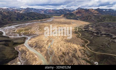 Ein geflochtenes Flusssystem fließt durch ein sandiges Tal mit zerklüfteten, bunten Bergen. Schneebesen säumen weit entfernte Gipfel im isländischen Hochland. Stockfoto