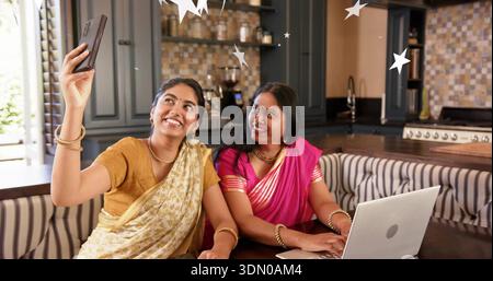Lächelnde Tochter, die ein Smartphone hält, Mutter sieht in Saris in der Küchenecke den Laptop Stockfoto