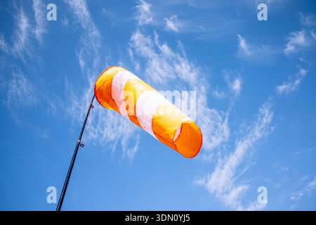 Orange-weiß gestreifte Windsocke, die die Windrichtung am Pol vor einem klaren blauen Himmel mit hellen Wolken anzeigt, Konzept der Wetterüberwachung Stockfoto