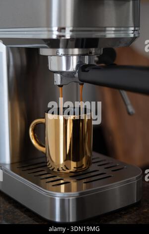 Nahaufnahme des Kaffees, der aus einer silbernen Kaffeemaschine zu Hause in ein vertikales Foto einer goldenen Tasse gegossen wird Stockfoto