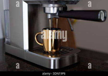 Frisch gebrühter Kaffee, der aus einer silbernen Espressomaschine in eine goldene Tasse fließt – warm, einladend und perfekt am Morgen Stockfoto