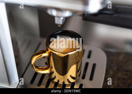 Frisch gebrühter Kaffee in einer goldenen Tasse aus einer silbernen Kaffeemaschine – cremiger Schaum, dampfend, bereit zum Schlucken, gemütlicher und eleganter Morgenkaffee Stockfoto