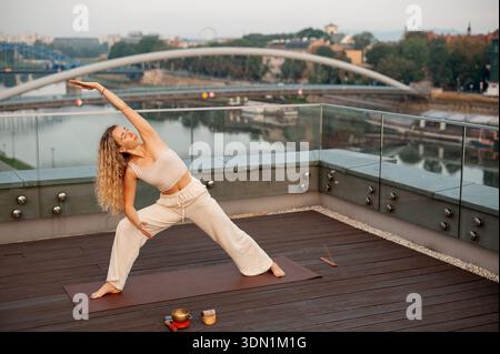 Eine Frau mit lockigen blonden Haaren spielt eine Yoga-Pose auf einer hölzernen Terrasse mit Blick auf einen Stadtfluss und eine Brücke Stockfoto