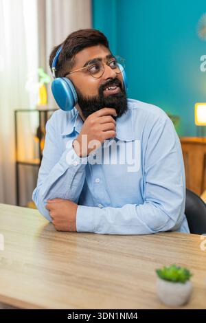 Indischer Mann sitzt zu Hause glücklicher Ausdruck, der Kopfhörer trägt, schwingt sanft tanzen und genießt ruhige, sanfte Musik. Arabian an der Rezeption hält Rhythmus Lächeln fühlt sich friedliche Stimmung und genießt Melodiemomente tief. Stockfoto