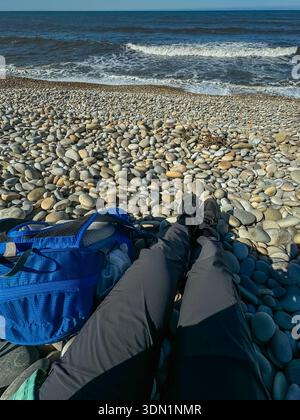 Der Standpunkt des Alleinreisenden, der am Kiesstrand mit Rucksack und Meereswellen vor sich liegt. Pause während der Wanderung auf dem Jakobsweg, langsame Fahrt Stockfoto