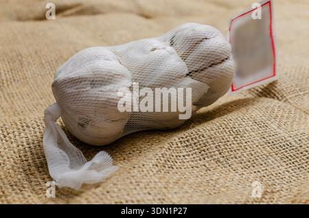 Eine Nahaufnahme von drei Bio-Knoblauchzwiebeln, verpackt in einem weißen Plastiknetz, auf einem rustikalen hessischen Jutestoff. Stockfoto