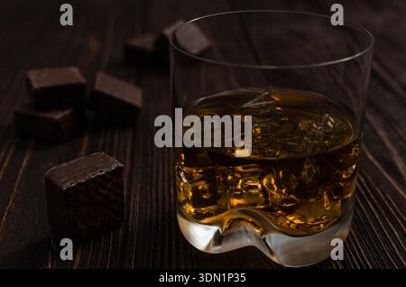 Nahaufnahme eines Glases bernsteinfarbenen Whiskeys mit Eiswürfeln, begleitet von dunklen Schokoladenpralinen auf rustikalem Holzhintergrund. Stockfoto