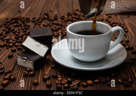 Heißer Kaffee wird aus einem Glastopf in eine weiße Keramikschale gegossen, begleitet von Milchschokolade und verstreuten gerösteten Kaffeebohnen Stockfoto