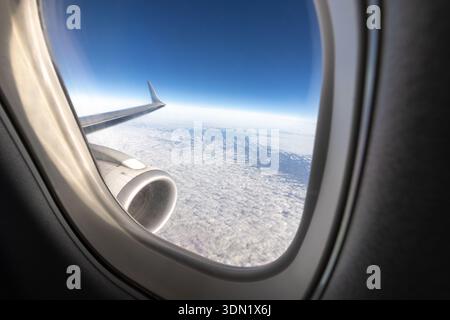 Blick auf einen Flugzeugflügel und -Motor über einem Wolkenmeer durch ein Flugzeugfenster in Reiseflughöhe unter tiefblauem Himmel. Thema Reise und Luftfahrt Stockfoto