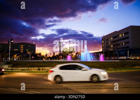 Eine dynamische Langzeitaufnahme eines weißen Autos, das vor einem bunten beleuchteten Brunnen und dem Benidorm Hofgebäude in der Dämmerung vorbeifährt. Stockfoto