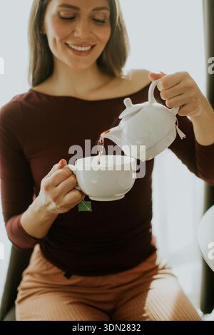 Eine Frau lächelt, als sie Tee aus einer Teekanne in eine Tasse gießt. Sie sitzt in einem hellen Raum mit Sonnenlicht durch ein Fenster. Die Atmosphäre s Stockfoto