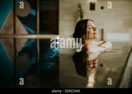 Die Frau genießt ihre Zeit in einem Hallenbad. Sie lehnt sich mit geschlossenen Augen zurück, und das Wasser reflektiert das Kunstwerk in der Nähe. Die Einstellung hat Stockfoto