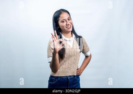 Fröhliche indonesische Frau, die mit einem Lächeln eine gute Handgeste zeigt, die Zufriedenheit, Zustimmung und Exzellenz auf weißem Hintergrund repräsentiert. Stockfoto