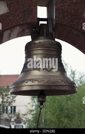Eine Nahaufnahme einer großen, verzierten Bronzeglocke mit einem geprägten Emblem, die in einem polierten roten Granitbogen hängt. Der Hintergrund ist leicht verschwommen. Sombe Stockfoto