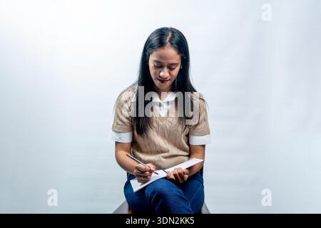 Ruhige junge indonesische Frau in lässiger, eleganter Kleidung, die in einem Notizbuch schreibt. Konzept des Studiums, Journals oder Planens auf einem sauberen weißen Hintergrund. Stockfoto