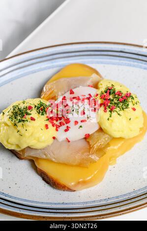 Toasten Sie mit Fisch und pochiertem Ei auf einer ovalen Platte neben einer Tasse Kaffee. Frühstück im Hotel mit einem heißen Sandwich und schwarzem Kaffee in einer Schüssel im t Stockfoto