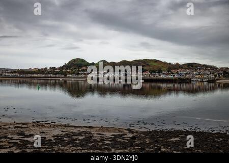 Ein stimmungsvoller Blick über die Mündung von Conwy bei Ebbe auf die Stadt Deganwy mit den Kamelhügeln der Burg an einem stimmungsvollen grauen Tag, Conway, Wales. Stockfoto