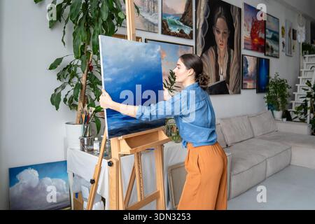 Fokussierte Künstlerin, die an der Malerei in der Galerie für Loft-Kunst arbeitet und fertige Kunstwerke an der Staffelei anpasst. Stockfoto
