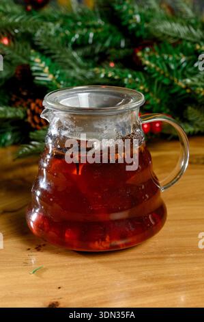 Teekanne aus Glas mit heißem Beerentee auf einem Holztisch. Gebrühter Früchtetee mit Gewürzen in einer transparenten Teekanne auf einem Tisch im Silvesterstil Stockfoto