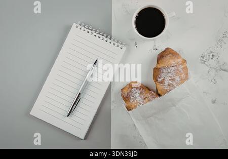 Eine ruhige Einrichtung am Morgen mit einer Tasse Kaffee, zwei Croissants, einem Notizblock und einem Stift auf einer Marmoroberfläche. Stockfoto