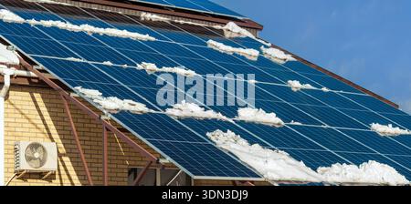 Sonnenkollektoren, die an einem Wintertag mit Schnee bedeckt sind, auf einem Dach vor blauem Himmel. Die Paneele liefern auch bei winterlichem Wetter saubere, erneuerbare Energie. Stockfoto