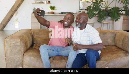 Lächelnde Männer in rosa T-Shirt, gestreiftes Hemd, die Selfie mit Telefon auf brauner Couch machen und Getränke halten Stockfoto