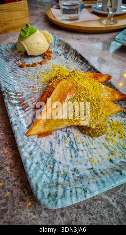 Gourmet-Dessertpräsentation mit knusprigen Baklava-Dreiecken, garniert mit zerstoßenen Pistazien, serviert mit einer Schaufel Vanilleeis und Minze auf einer Textur Stockfoto