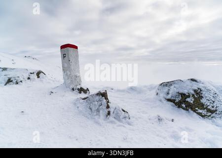 Einsamer Grenzposten, der von einem eisigen Bergkamm aufsteigt, bedeckt mit Frost und Schnee, in starkem Kontrast zu weißem Gelände, während dicker Nebel und Wolken schweben Stockfoto