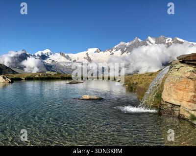 Malerischer kristallklarer Bergsee mit kleinem Wasserfall, umgeben von beeindruckenden Bergbergen, Kreuzboden, SaaS Fee, Wallis, Schweiz Stockfoto