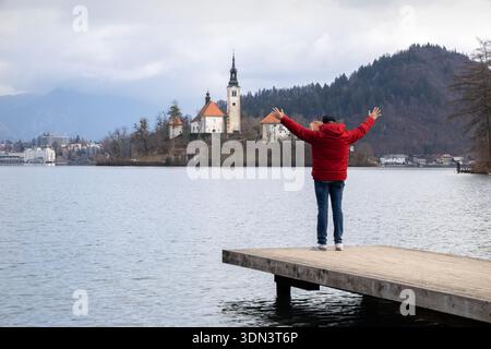 Reisende in roter Jacke auf Holzsteg mit erhobenen Armen und Blick auf die Kirche der Insel Bleder See und die Berge in Slowenien Stockfoto