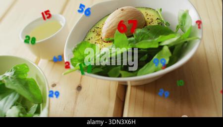 Weiße Schüssel mit halbierter Avocado mit Grube, Grünzeug auf Holztisch, numerische Überlagerungen Stockfoto