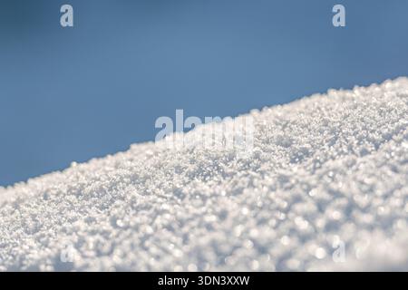 Aus nächster Nähe sehen Sie glitzernde Schneekristalle, die sanft von Sonnenlicht beleuchtet werden und einen hellen, strukturierten Winterhintergrund mit einem ruhigen und minimalistischen Ambiente schaffen Stockfoto