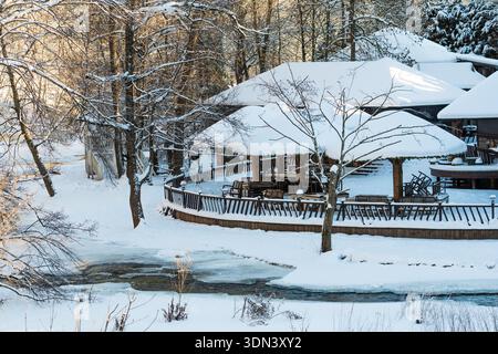 Schneebedeckte hölzerne Restaurantterrasse an einem teilweise gefrorenen Fluss, umgeben von Winterbäumen, schafft eine ruhige saisonale Szene in einem Naturpark Stockfoto