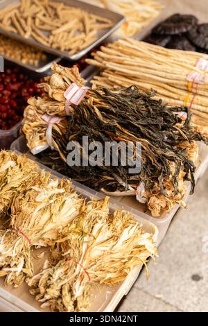 Getrocknete Kräuter und Wurzeln werden auf Tischen auf einem lokalen Markt ausgestellt. Kunden erkunden die Auswahl der Pflanzen. Der Tag ist warm und hell, typisch für AUT Stockfoto