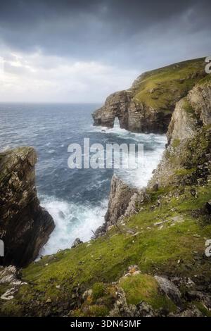 Der Blick auf dramatische Klippen trifft auf das turbulente Meer unter einem stürmischen Himmel, während Wellen gegen die zerklüftete Küste von Dunfanaghy, County Donegal, Irland, krachen. Stockfoto