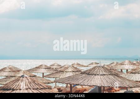 Strohschirme am Meer unter hellem Himmel, sanfter Hintergrund mit glitzerndem Wasser und Bergen in der Ferne, ruhig Stockfoto