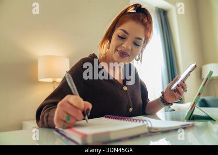 Die junge Frau sitzt zu Hause an einem Tisch, schreibt in ein Notizbuch, während sie ihr Smartphone überprüft. Entspannte tägliche Routine, persönliche Organisation und Balance Be Stockfoto