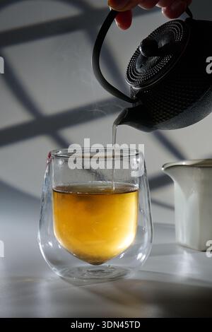 Gießen Sie von Hand heißen Tee aus einer traditionellen gusseisernen Teekanne in ein doppelwandiges Glas, wobei Dampf aus dem goldenen Getränk steigt Stockfoto