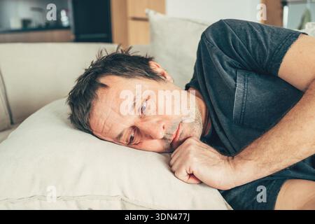 Depressiver reifer Mann, der mit traurigem Ausdruck auf der Couch liegt und mit emotionalen Schmerzen, Einsamkeit oder psychischen Gesundheitsproblemen in einem ruhigen Wohnraum kämpft. Stockfoto