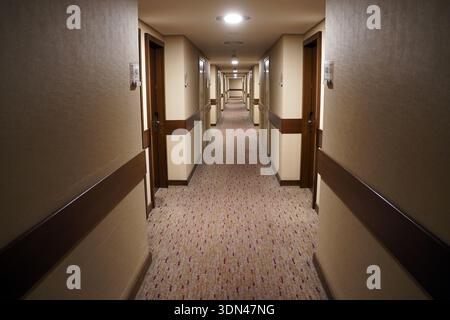 Ein langer Hotelflur mit Teppichboden erstreckt sich in die Ferne mit zahlreichen identischen Holztüren, die beide Seiten unter Deckeneinbauleuchten säumen Stockfoto