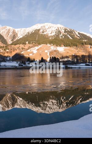 Schneebedeckte Berge und alpines Dorf, das sich im Winter auf einem teilweise gefrorenen See spiegelt Stockfoto