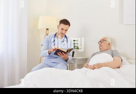 Pflegende männliche Krankenschwester, die bei einem älteren Patienten im Bett sitzt, ihm Buch liest, Unterstützung in der Klinik leistet. Stockfoto