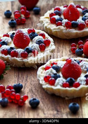 Mini-Frucht-Törtchen, die mit Schlagsahne, Himbeeren, Blaubeeren und roten Johannisbeeren gekrönt sind, auf einer rustikalen Holzoberfläche angeordnet. Stockfoto