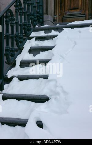Holztreppen sind mit Schnee bedeckt, der sich auf dem Boden stapelt und zu einer dunklen Tür und einem Metallgeländer führt. Stockfoto