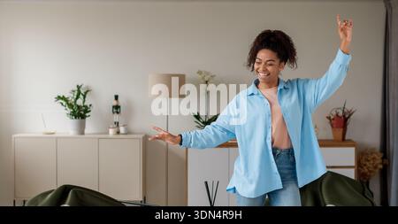 Frau tanzt glücklich im modernen Wohnzimmer mit hellblauem Hemd und Jeans Stockfoto