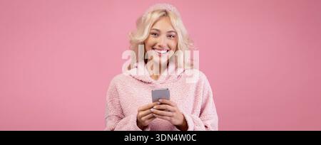 Lächelnde Frau mit Telefon in gemütlicher Kleidung steht vor rosa Hintergrund Stockfoto
