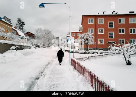 Eine Frau läuft nach einem Schneesturm eine verschneite Straße vor einem roten Gebäude entlang. Die Straße ist mit Schnee bedeckt und die Frau trägt einen schwarzen Mantel. Stockfoto