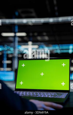 Green-Screen-Laptop, der vom Administrator des Rechenzentrums zur Inspektion der Infrastruktur und zur Aktualisierung von Netzwerksystemen verwendet wird. Nahaufnahme eines Mitarbeiters, der ein Modell-Notebook verwendet, um einen stabilen Server-Hub-Betrieb zu gewährleisten, und die Steuerung von Rigs Stockfoto