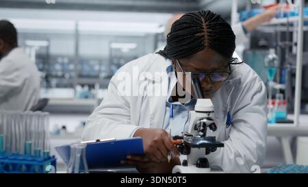 Wissenschaftlerin mit optischem Mikroskop zur Untersuchung von Mikroorganismen im Labor, innovativ. afroamerikanerin, die durch detaillierte biologische Forschung zur wissenschaftlichen Gemeinschaft beiträgt, Kamera B Stockfoto